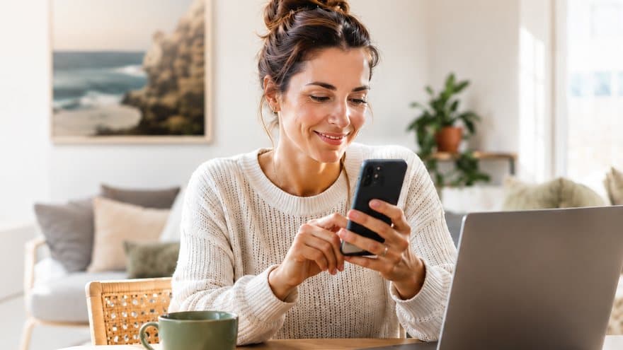 A woman smiling at her phone beside her laptop at home